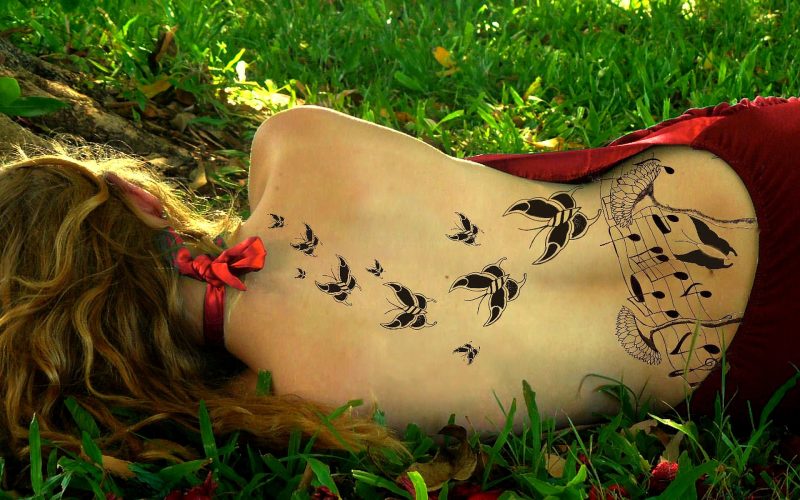 Chica tumbada en la hierba de espaldas a la cámara con la espalda al descubierto y toda ella tatuada