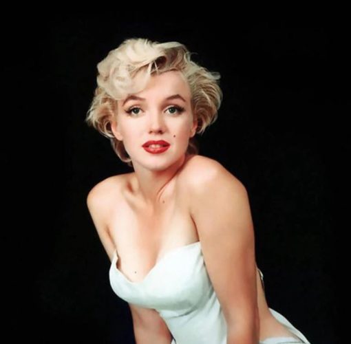 Marilyn Monroe mirando a la cámara directamente. Va vestida con un vestido blanco escotado sobre fondo negro.