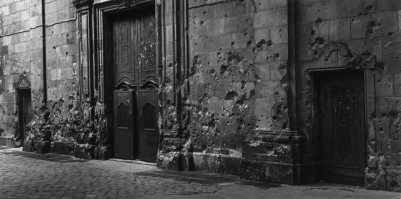 Foto en blanco y negro de una puerta grande antigua y una pared llena de agujeros (quizá por impactos de bala)