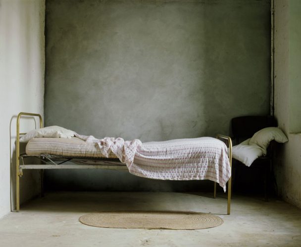Foto de una cama muy simple vista desde un lado, con una silla al fondo y la almohada encima