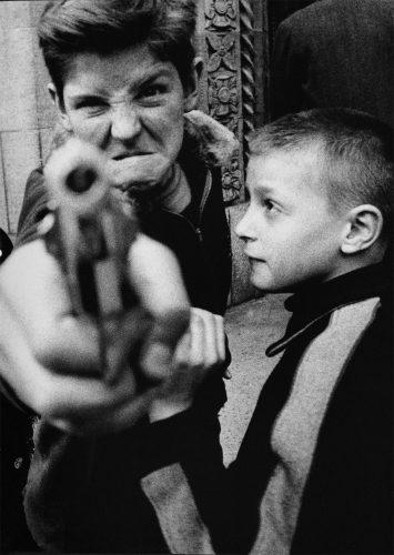 Foto en blanco y negro de dos niños, uno de ellos apunta con una pistola directamente a la cámara.