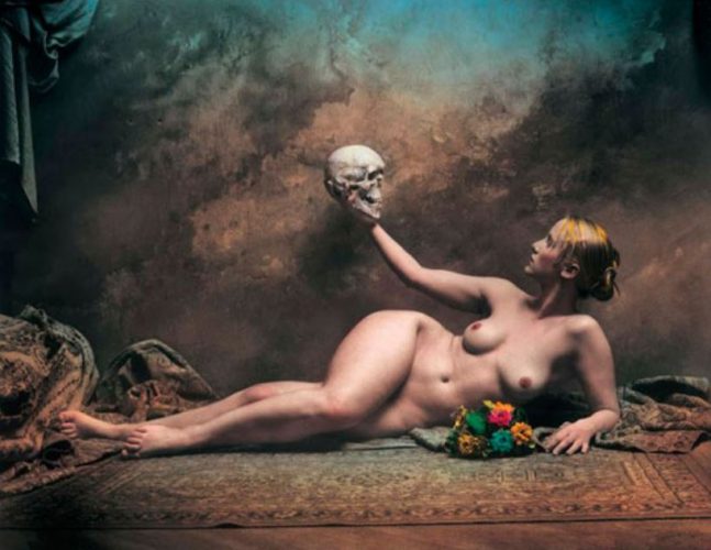 Foto artística de una mujer desnuda recostada en el suelo mientras con su mano derecha sujeta una calavera humana