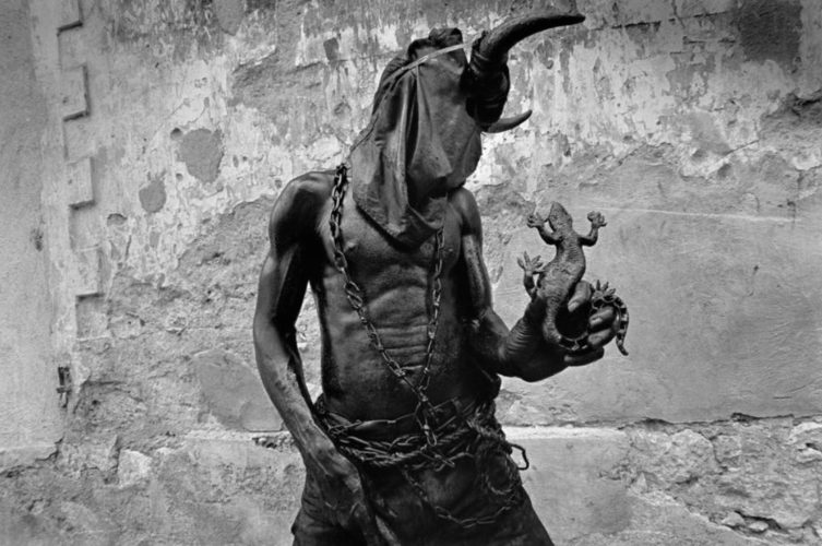 Foto tribal en blanco y negro donde aparece un hombre con la cabeza tapada y encima unos cuernos, mientras sujeta un lagarto con una mano