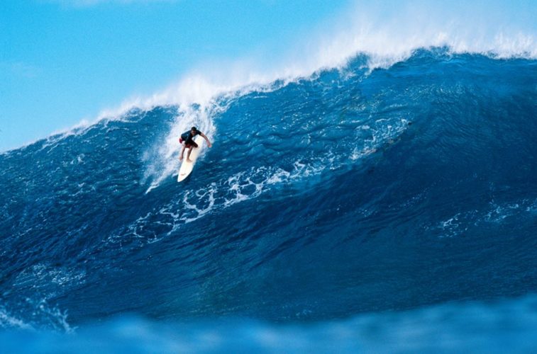 Foto de un hombre haciendo surf sobre unas olas azules gigantescas.