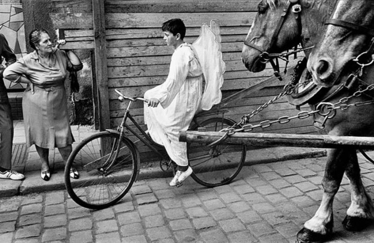 Foto en blanco y negro de un niño disfrazado de ángel con alas paseando en una bicicleta mientras una mujer le mira