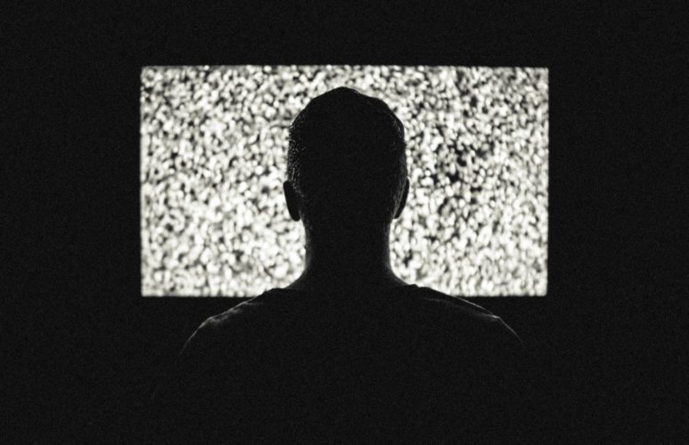 Un hombre en la obscuridad mirando la TV en la cual se puede ver ruido, nieve como en Poltergeist