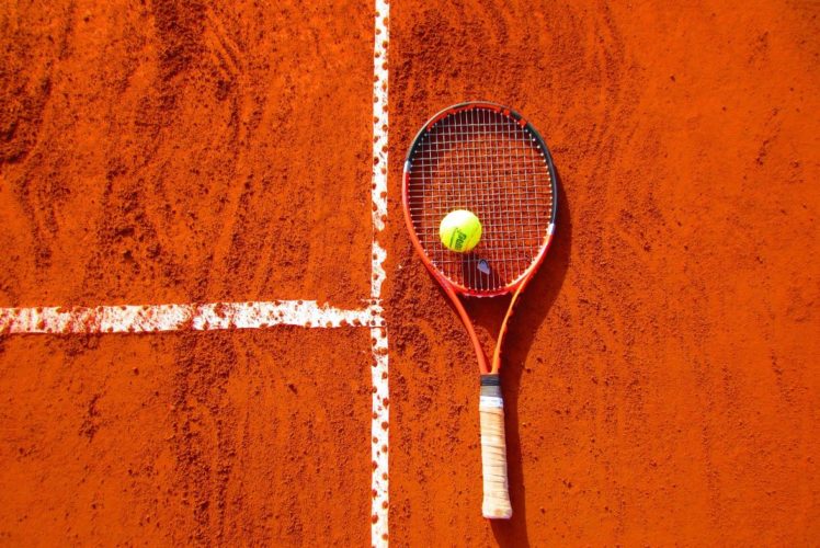 En la imagen una raqueta de tenis con una pelota sobre ella en el suelo de una pista de tierra batida
