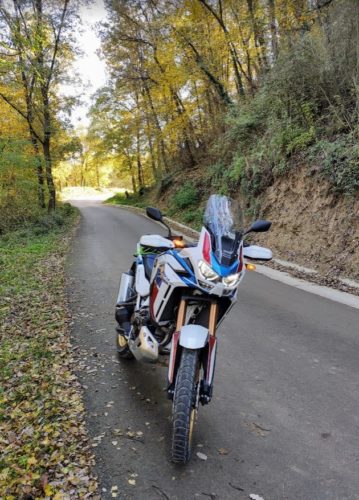 Se ve la moto parada en el lateral de una calzada con sus márgenes recubiertos de hojas en el mes de otoño