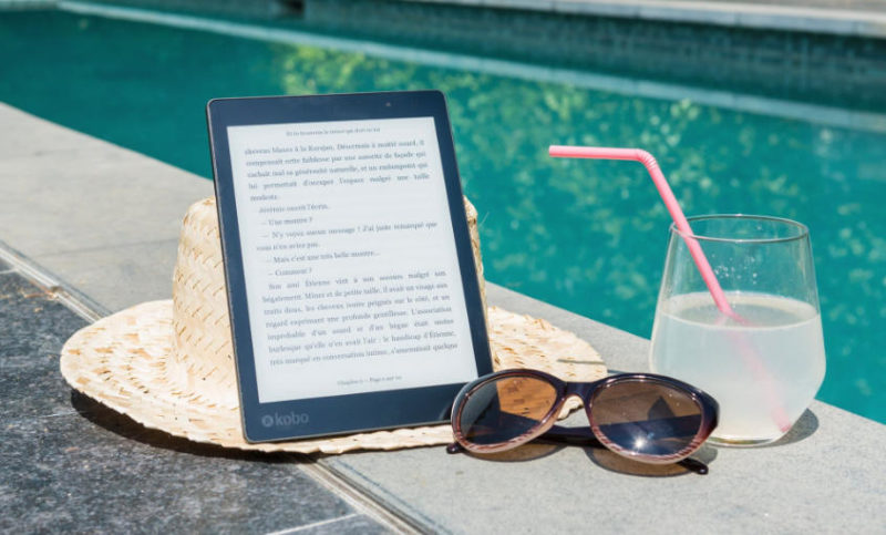 En la foto un ebook, unas gafas de sol y un cocktail en primer plano, al fondo una piscina